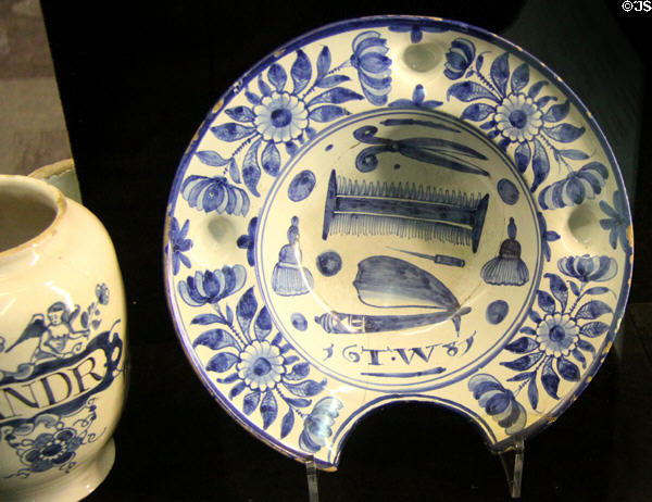 Tin-glazed earthenware barber's bowl painted with comb, scissors & other tools of profession & cutout to fit client's neck (1681) by TW of London at Potteries Museum & Art Gallery. Hanley, Stoke-on-Trent, England.