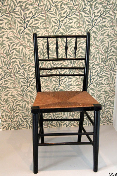 Arts & Crafts Sussex chair (mid 1860s) by Morris & Co against Willow Bough wallpaper (reproduction first designed 1887) by William Morris at Morris Gallery. London, United Kingdom.