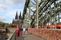 Pedestrians strolling across Hohenzollern Bridge with train crossing and multitude of love locks with Köln Cathedral beyond. Köln, Germany.