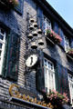 Shingled building adorned with flowers, decorative shutters & rows of bells. Monschau, Germany.